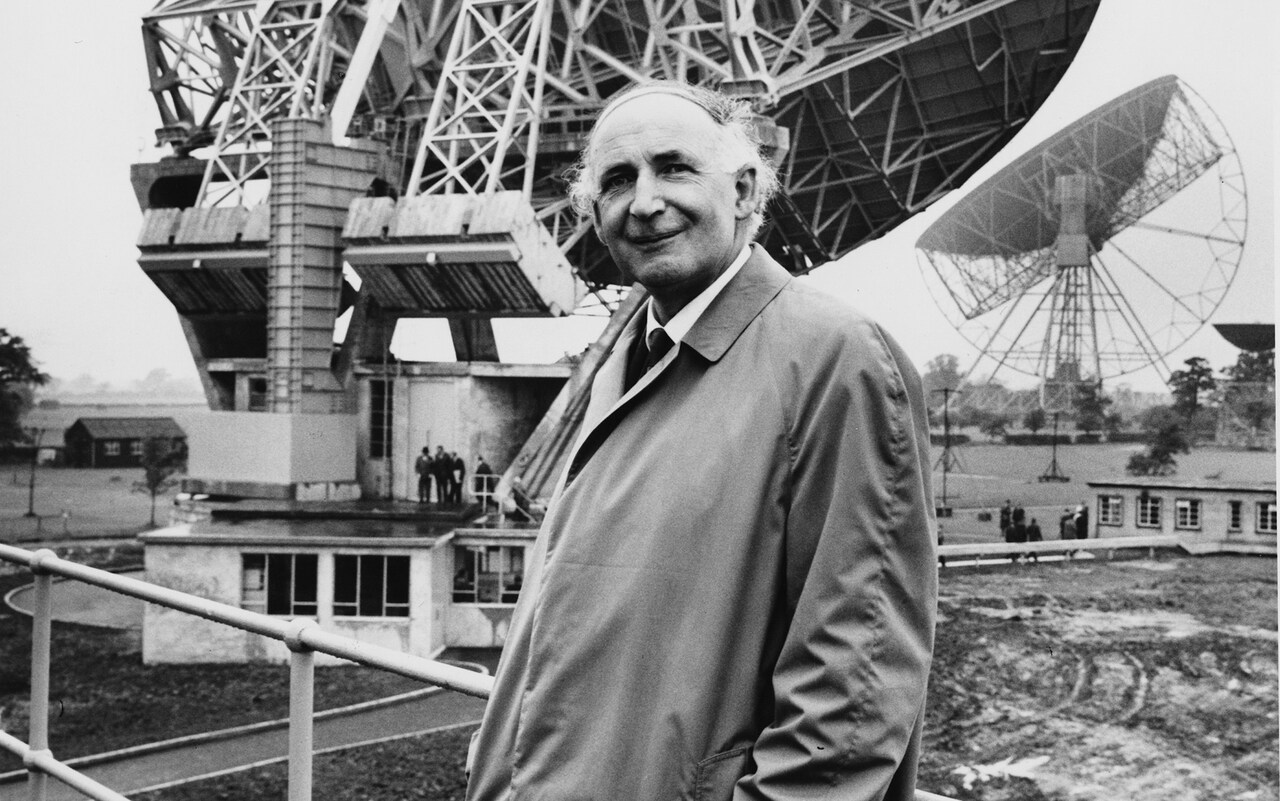 A black and white photo of Sir Bernard Lovell standing in front of the Lovell Telescope at Jodrell Bank, Cheshire